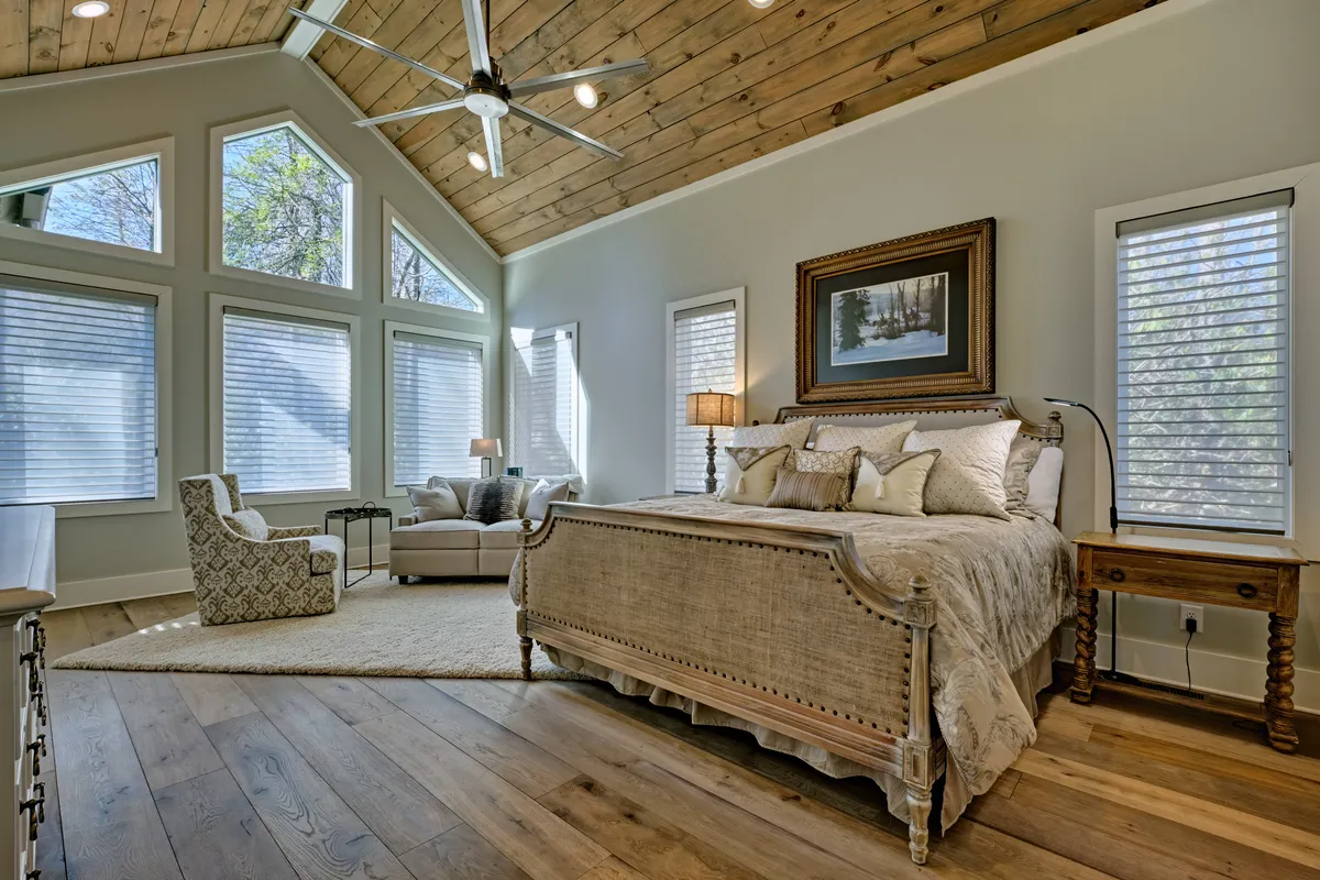 A luxurious bedroom featuring a large bed with decorative pillows, expansive windows allowing in natural light, a wooden ceiling, and a comfortable seating area. The layout exudes tranquility and comfort, perfectly suited for relaxation and privacy.