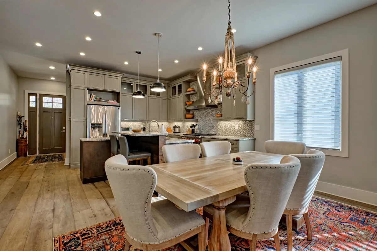 This scene features a spacious dining area with a large table under a chandelier, a kitchen island in the background, and decorative tilework. The arrangement reflects a place for shared meals and elegant entertaining, with finishes that suggest luxury and comfort.