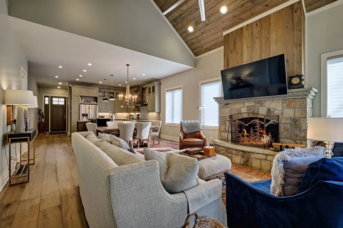 This image showcases a spacious and inviting living area featuring a cozy fireplace, comfortable sofa, elegant dining table, and chandelier, all positioned to create a warm gathering space. Large windows (not specified but often present in such rooms) may offer glimpses of the outdoors, subtly referencing a connection to nature.