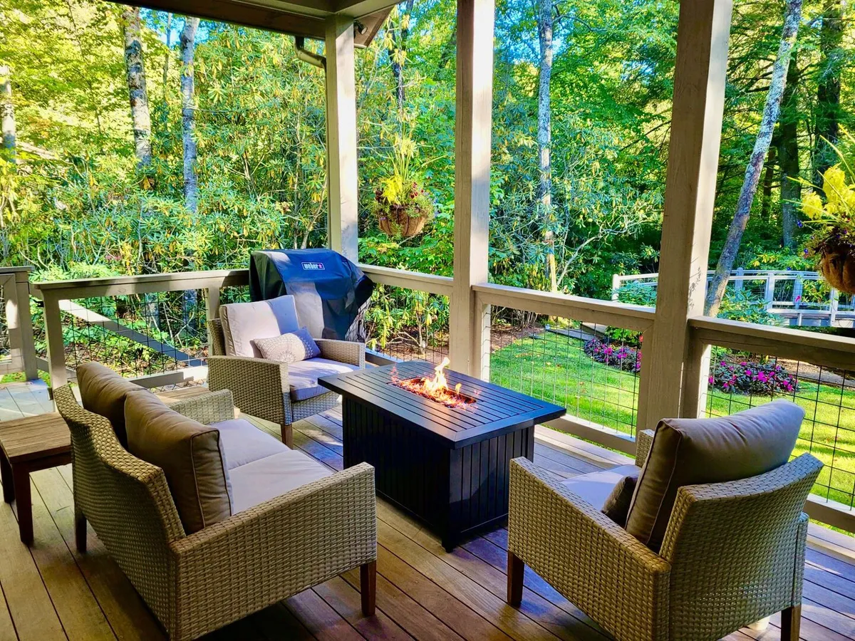The image showcases an inviting outdoor area surrounded by lush greenery, featuring a cozy fire pit with comfortable seating, a grill for barbecues, and ample space for gathering with friends and family. The setting feels intimate and perfect for enjoying the tranquil mountain air or making memories under the stars.