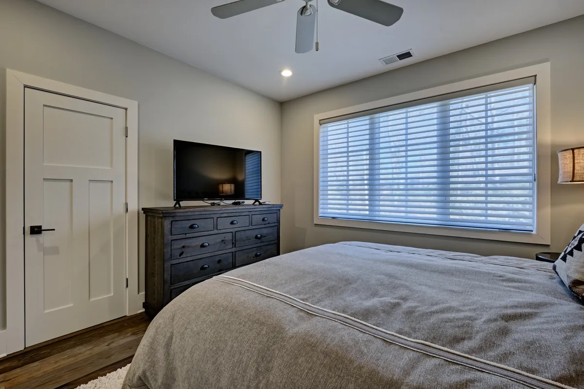 A restful bedroom with a plush bed, textured bedding, a television, and a large window with blinds. The room is styled with soothing colors and modern furnishings, offering both comfort and entertainment for guests looking to relax after busy days.