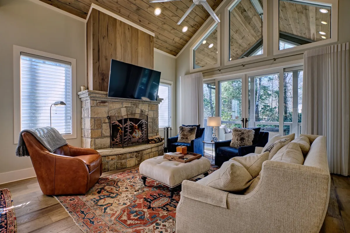 This image spotlights a cozy living room with a prominent stone fireplace, large windows inviting natural light, comfortable designer seating, a decorative rug, and a flat-screen TV. The arrangement suggests a welcoming environment for relaxation and social gatherings.